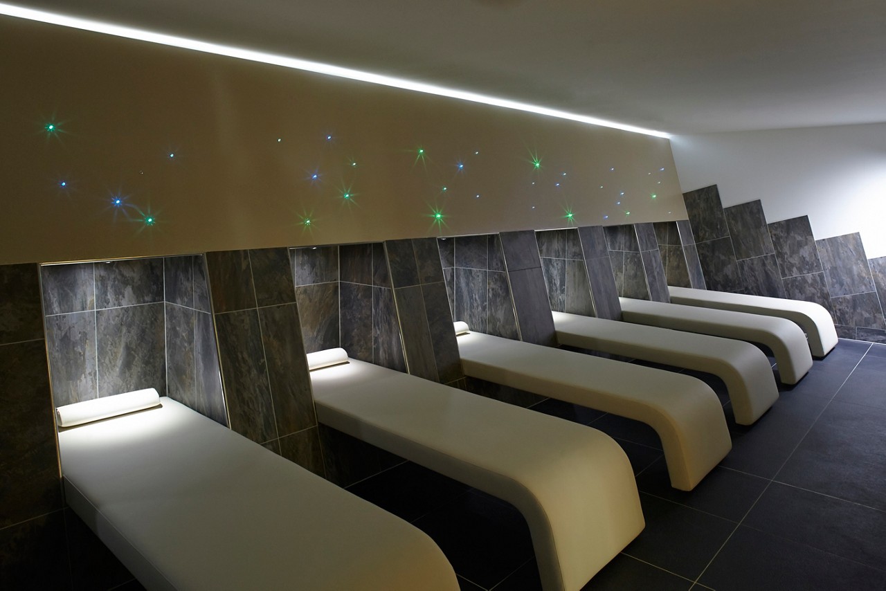 Curved lounge beds align in a row, inviting rest, within a dim, tiled spa room featuring underlit headrests, a ceiling light strip, and scattered green and blue star-like wall lights.